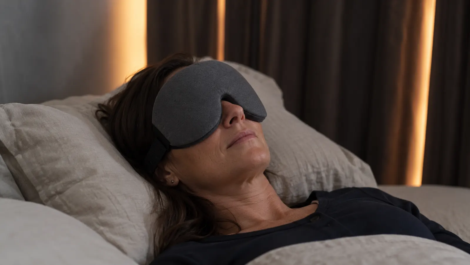 Woman lying in bed in a dark bedroom wearing a contoured sleep mask, early morning light visible at curtain edges
