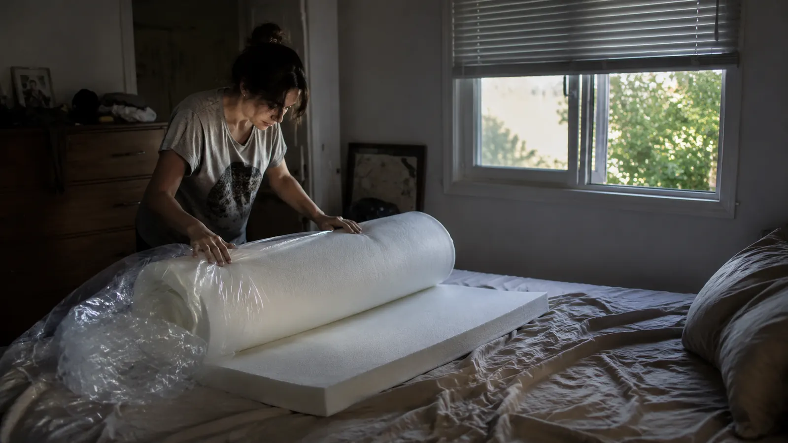 Rolled memory foam mattress topper partially unrolled on a bed in a dimly lit bedroom, suggesting the product just arrived and is off-gassing
