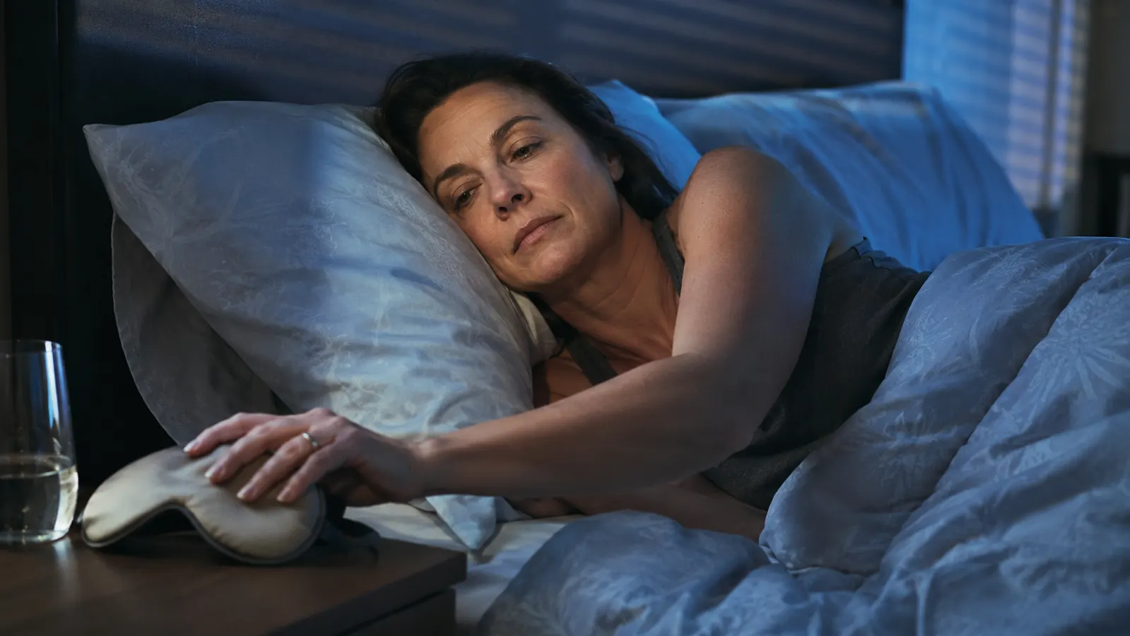Woman lying awake in a dim bedroom, city glow visible through window blinds, reaching for a sleep mask on the nightstand
