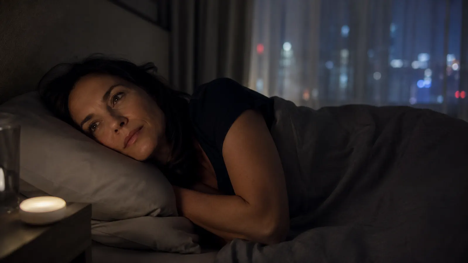 Woman lying awake in bed at night, small white noise machine visible on the nightstand, looking thoughtful rather than distressed
